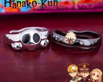 May include: Two silver rings, one with a black and white skull design and the other with a gold flower design. The rings are on a dark red surface. The text "Toilet-bound Hanako-Kun" is visible in the background.