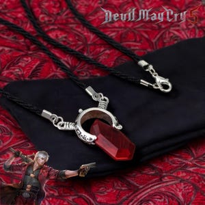 Może przedstawiać: Czarny naszyjnik ze sznurka ze srebrnym wisiorkiem z dużym, fasetowanym, czerwonym kryształem. Wisiorek ma kształt podkowy z wygrawerowanymi detalami. Obraz zawiera również tekst "Devil May Cry 5".