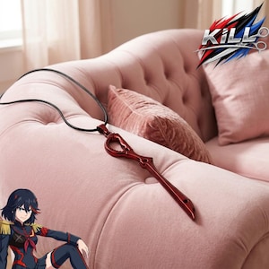 Op de afbeelding: Een rode schaarvormige hangerketting met een zwart koord. De hanger is een gestileerd ontwerp, waarschijnlijk uit een anime- of mangaserie. De ketting wordt getoond op een roze, gecapitonneerde bank met kussens.