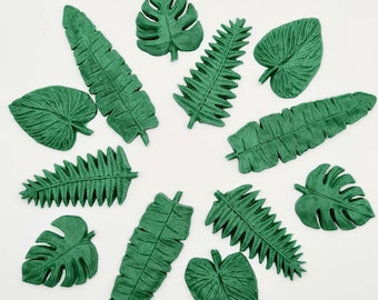 May include: A collection of green, textured artificial leaves in various shapes and sizes, including monstera, fern, and banana leaf designs. The leaves are arranged on a white surface, creating a tropical theme.