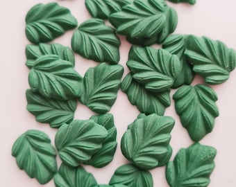 May include: A collection of small, green leaf-shaped decorations. Each leaf has detailed veining and a slightly textured surface. The leaves are scattered on a light pink background, creating a natural, decorative theme.
