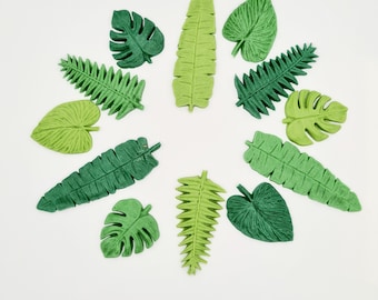 May include: An assortment of artificial leaves in various shades of green, arranged in a circular pattern. The leaves include different shapes and sizes, such as fern, banana, and monstera leaves. The leaves are set against a white background.