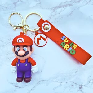 May include: A Super Mario keychain featuring a red-capped Mario figure with a mustache, red shirt, and purple overalls. The keychain includes a red strap with the words "SUPER MARIO" and a gold-colored keyring.