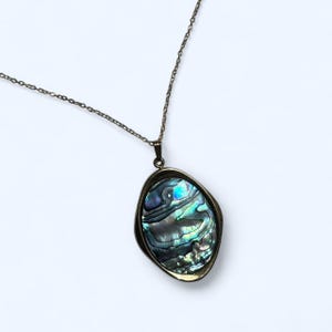 Puede incluir: Collar con colgante de concha de abulón. El colgante presenta una concha de abulón pulida e iridiscente en un bisel de tono bronce. La concha muestra tonos de azul, verde y plata. La cadena del collar también es de tono bronce.