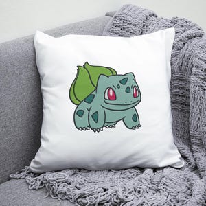 May include: White pillow with a green and blue cartoon image of a Pokemon character, Bulbasaur, with red eyes. The character is on a white background.