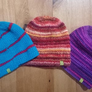 以下が含まれることがあります： 青、赤、紫の色合いのニット帽が3つ。青い帽子には赤いストライプがあり、真ん中の帽子は赤、オレンジ、黄色の混合色で、紫の帽子にはポンポンが付いています。各帽子には小さな緑色のタグが付いています。