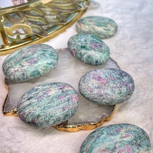 May include: Six smooth, oval-shaped polished stones featuring a mottled pattern of teal, green, and white with distinct inclusions of vibrant magenta and deep pink. The stones are arranged on a white, irregular-shaped slab with a metallic gold-toned edge.