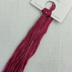 May include: Close-up of raspberry-coloured embroidery floss. The floss is hand-dyed and comes with a tag that reads "Raspberry" and "75m/8.2 yards". The background is a white, textured fabric.