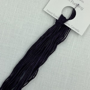 Puede incluir: Un ovillo de hilo de bordar morado oscuro etiquetado como "Plum Pudding", con el texto "Hand Dyed" y "75m/8.2 yards". El hilo está anudado y descansa sobre un fondo de lino blanco.