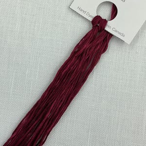 May include: A skein of hand-dyed Sangria embroidery thread. The thread is a rich burgundy hue and is attached to a white label. The label reads "Sangria" and "7.5m/8.2yards". The label also includes the text "Hand Dyed".