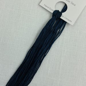 Può includere: Matassa di filo da ricamo tinto a mano di colore blu intenso, etichettata "Deep Blue Sea". L'etichetta indica 7,5 m di filo. Il filo è annodato e appoggiato su uno sfondo bianco strutturato.