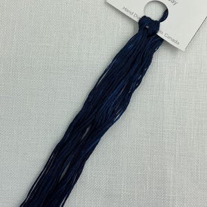 Peut inclure: Écheveau de fil à broder teint à la main, bleu foncé, étiqueté "Blue Jay". L'étiquette indique 75 m de fil. Le fil est présenté sur un fond de tissu blanc texturé.
