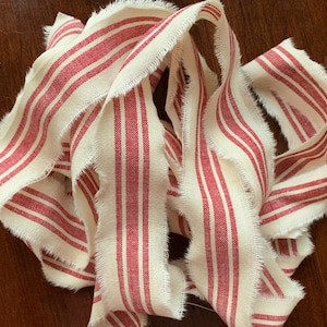 May include: A close-up of a red and white striped fabric ribbon with frayed edges. The ribbon is made of a natural linen material and has a rustic, farmhouse style.