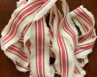 May include: A close-up of a red and white striped fabric ribbon with frayed edges. The ribbon is made of a natural linen material and has a rustic, farmhouse style.