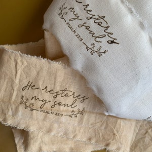 May include: Close-up of two fabric ribbons with frayed edges. The ribbons are cream and off-white. The text "He restores my soul, Psalm 23:3" is printed in brown script with floral accents. The ribbons are layered.