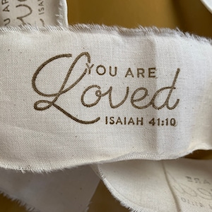 May include: A white fabric tag with frayed edges, featuring the text "You are Loved" in a cursive font and the scripture reference "Isaiah 41:10" in a smaller font.