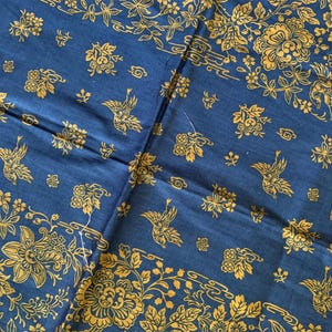 May include: A close-up of a blue fabric with a gold floral and bird pattern. The design includes stylized flowers, birds in flight, and decorative borders. The fabric is folded, showing the pattern's repeat.