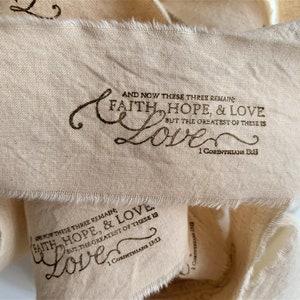 May include: A beige fabric with a brown printed quote: "And now these three remain: Faith, Hope, & Love. But the greatest of these is Love. 1 Corinthians 13:13"