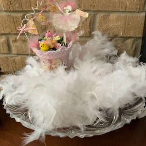 May include: A pink ballerina figurine with a white tutu and a pink hat, holding a sign that says "Joy". The figurine is surrounded by white feathers and small yellow chicks in a pink and white cup.