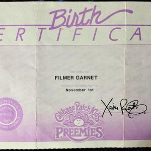 May include: A vintage Cabbage Patch Kids Preemies birth certificate. The certificate is light purple with the words "Birth Certificate" in a cursive font. It includes the name "Filmer Garnet" and the date "November 1st".