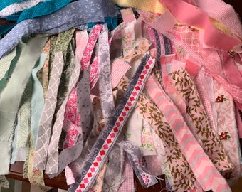 May include: A collection of fabric scraps in various shades of pink, green, gray, and blue. The fabric scraps are cut into long strips and have different patterns, including floral, geometric, and striped.