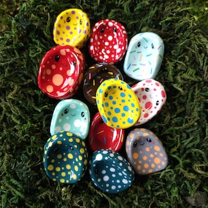 Op de afbeelding: Een verzameling kleurrijke, handgeschilderde keramische schaaltjes met lachende gezichten en diverse stip- en strooiselpatronen. De schaaltjes zijn in rood, geel, blauw en bruin, geplaatst op een groene mos achtergrond.