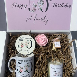 K&ouml;nnte beinhalten: Eine wei&szlig;e Geschenkbox mit rosa Deckel, die eine Tasse, eine Kerze, einen Untersetzer und eine rosa Rose enth&auml;lt. Die Tasse und die Kerze sind mit floralen Designs und dem Text "Happy Birthday 50 Mandy" verziert. Die Box ist mit braunem Papier gef&uuml;llt.