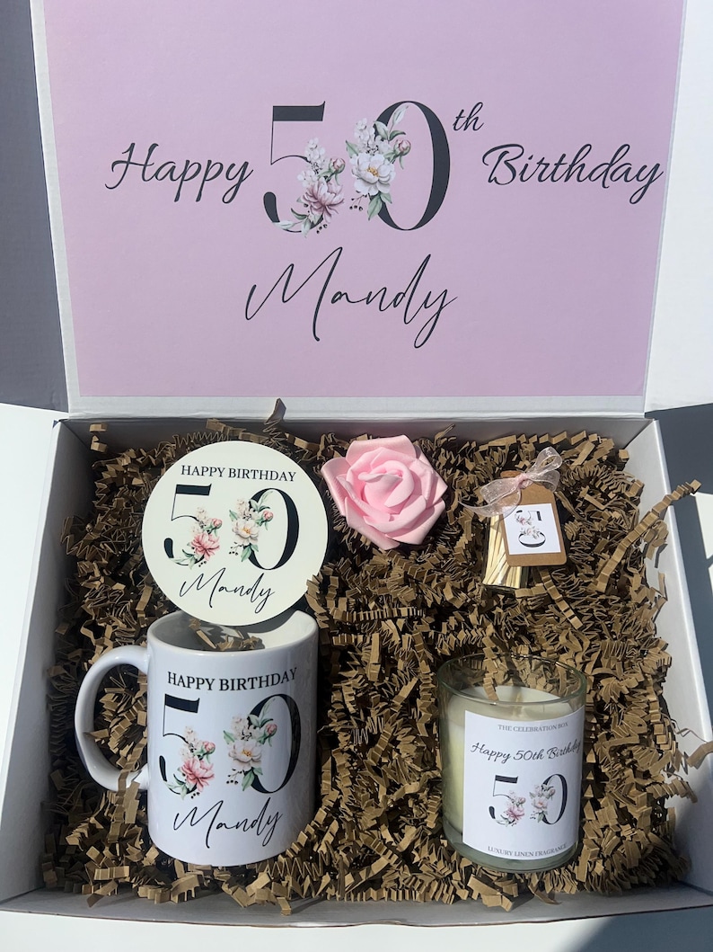 K&ouml;nnte beinhalten: Eine wei&szlig;e Geschenkbox mit rosa Deckel, die eine Tasse, eine Kerze, einen Untersetzer und eine rosa Rose enth&auml;lt. Die Tasse und die Kerze sind mit floralen Designs und dem Text "Happy Birthday 50 Mandy" verziert. Die Box ist mit braunem Papier gef&uuml;llt.
