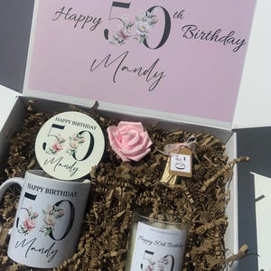 K&ouml;nnte beinhalten: Geschenkbox zum Geburtstag mit rosa Deckel, enth&auml;lt eine Tasse, einen Untersetzer, eine Kerze und eine rosa Rose. Die Artikel sind mit floralen Designs und dem Text "Happy 50th Birthday Mandy" verziert. Die Box ist mit Papier gef&uuml;llt.