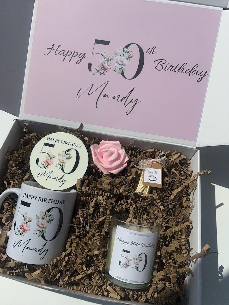 K&ouml;nnte beinhalten: Geschenkbox zum Geburtstag mit rosa Deckel, enth&auml;lt eine Tasse, einen Untersetzer, eine Kerze und eine rosa Rose. Die Artikel sind mit floralen Designs und dem Text "Happy 50th Birthday Mandy" verziert. Die Box ist mit Papier gef&uuml;llt.