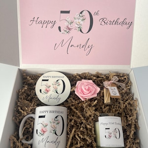 K&ouml;nnte beinhalten: Eine wei&szlig;e Geschenkbox mit rosa Innenraum, die eine Tasse, eine Kerze, einen Untersetzer und eine rosa Rose enth&auml;lt. Die Tasse, der Untersetzer und die Kerze sind mit floralen Designs und dem Text "Happy 50th Birthday Mandy" verziert. Der Deckel der Box lautet "Happy 50th Birthday Mandy".