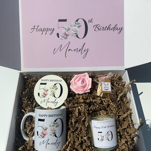 K&ouml;nnte beinhalten: Geschenkbox zum Geburtstag mit rosa Deckel, auf dem "Happy 50th Birthday Mandy" steht. Im Inneren befinden sich eine Tasse, ein Untersetzer, eine Kerze und eine rosa Rose. Tasse und Kerze tragen den Aufdruck "Happy Birthday 50 Mandy".