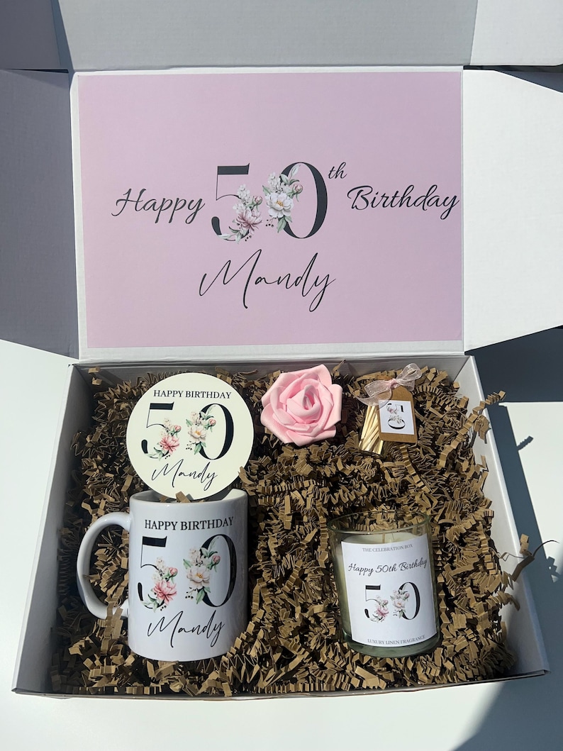 K&ouml;nnte beinhalten: Geschenkbox zum Geburtstag mit rosa Deckel, auf dem "Happy 50th Birthday Mandy" steht. Im Inneren befinden sich eine Tasse, ein Untersetzer, eine Kerze und eine rosa Rose. Tasse und Kerze tragen den Aufdruck "Happy Birthday 50 Mandy".