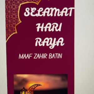 Op de afbeelding: Een rode en gouden kaart met de tekst "Selamat Hari Raya" en "Maaf Zahir Batin". De kaart heeft een afbeelding van een halve maan en een lantaarn tegen een zonsondergang achtergrond.