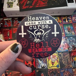 May include: A circular sticker with a dark background featuring a central winged figure, two inverted crosses, and two pentagrams. The text reads: Heaven came with a curse. Hell set me free. The design uses white and red lettering on a dark blue surface.