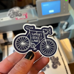 May include: A dark blue die-cut sticker shaped like a bicycle with white outlines and text that reads I SUPPORT BIKE LANES. The sticker features a detailed frame, two wheels with spokes, a seat, and handlebars, presented in a clean graphic style.