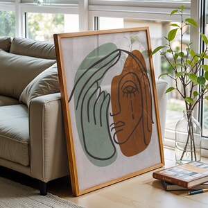 Può includere: Una stampa artistica astratta incorniciata con un disegno a tratto minimalista di un volto e una mano nei toni del marrone, verde e nero. L'opera d'arte è esposta su uno sfondo neutro e incorniciata in legno chiaro. La stampa è appoggiata a un divano beige.