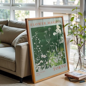 Può includere: Un poster incorniciato con cornice in legno è appoggiato a una finestra. Il poster presenta il testo "FLOWER MARKET TOKYO" in rosso e bianco, con uno sfondo verde e illustrazioni floreali bianche. Un divano beige e un vaso di vetro con vegetazione sono visibili.
