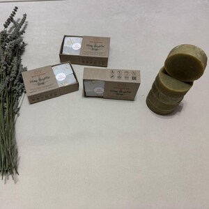 Pode incluir: Uma disposição de produtos naturais à base de cânhamo. A imagem apresenta três caixas de sabão, uma pilha de sabonetes redondos e um ramo de lavanda seca. O sabão é de cor verde claro e as caixas são castanhas.