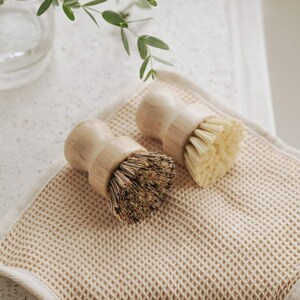 May include: Two wooden-handled scrub brushes with natural bristles. The brushes are on a beige waffle-weave kitchen towel.