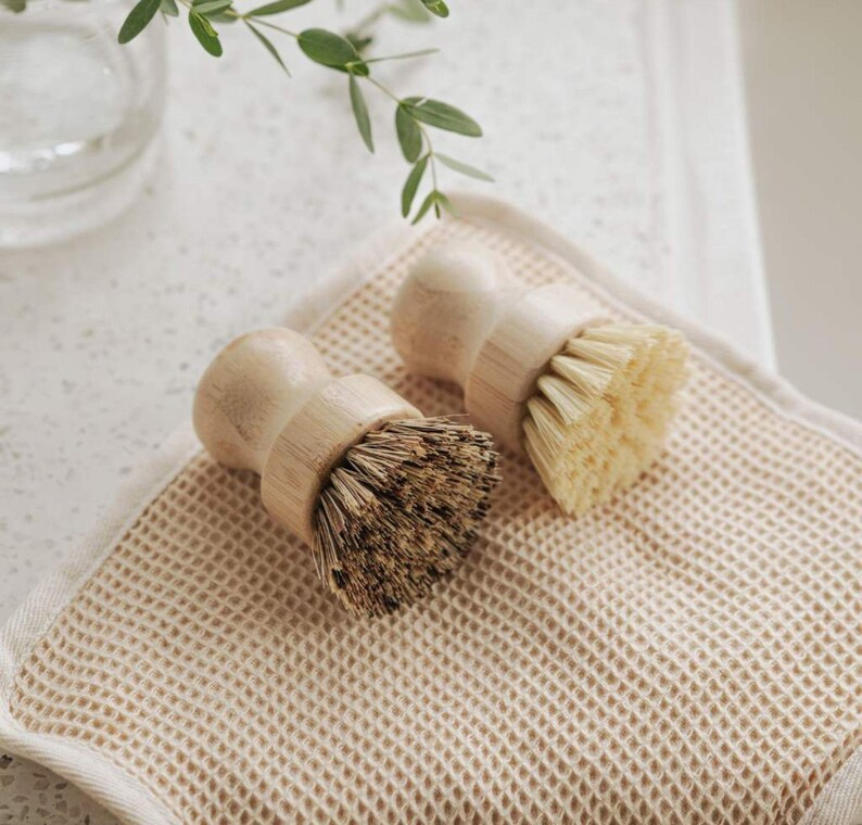 May include: Two wooden-handled scrub brushes with natural bristles. The brushes are on a beige waffle-weave kitchen towel.