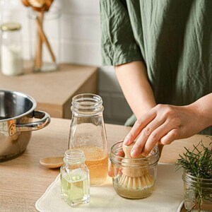 May include: A close-up of a glass jar with a wooden brush inside. The jar is sitting on a white surface with a clear glass bottle and a wooden spoon. The jar is filled with a clear liquid.