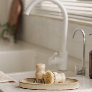 May include: Two wooden handled brushes with natural bristles are displayed on a light beige oval ceramic dish. The dish is sitting on a white countertop in a kitchen sink.