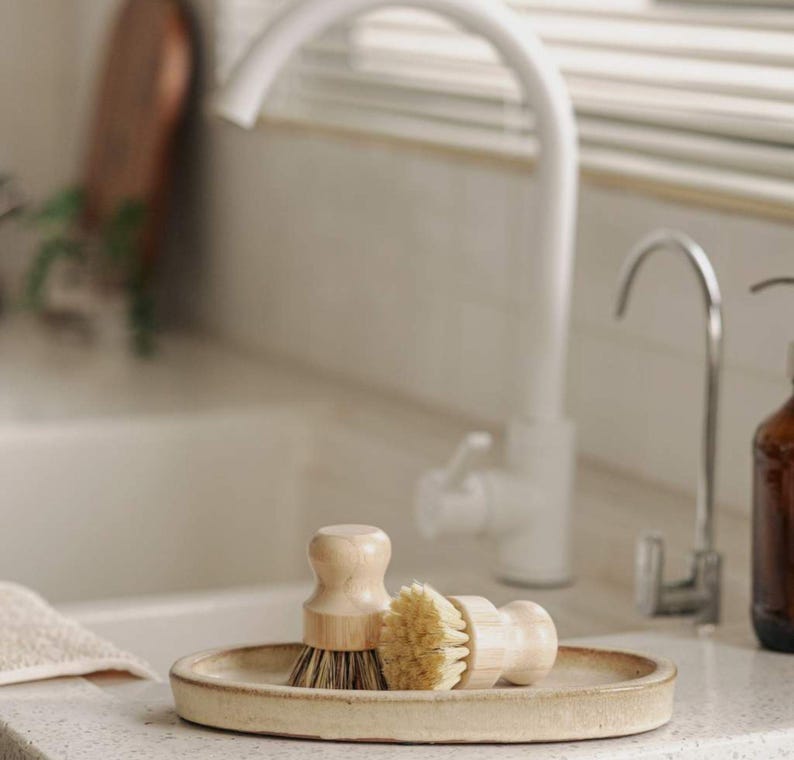 May include: Two wooden handled brushes with natural bristles are displayed on a light beige oval ceramic dish. The dish is sitting on a white countertop in a kitchen sink.