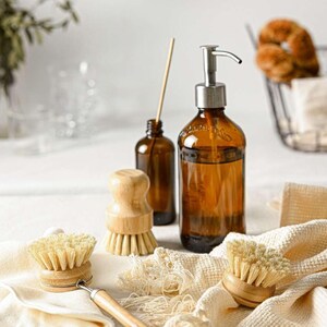 May include: A set of cleaning supplies including a brown glass bottle with a pump dispenser, a brown glass bottle with a wooden stopper, and three wooden-handled brushes with natural bristles.