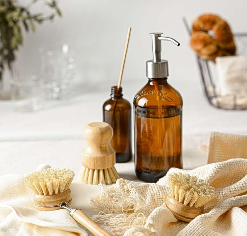 May include: A set of cleaning supplies including a brown glass bottle with a pump dispenser, a brown glass bottle with a wooden stopper, and three wooden-handled brushes with natural bristles.