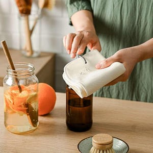 May include: A person is using a white cotton dishcloth to wipe a glass bottle with a brown liquid inside. The bottle is sitting on a wooden surface next to a glass jar with orange peels and water.