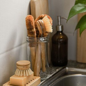 May include: A glass jar filled with four brown and beige dish brushes with wooden handles. The jar is sitting on a countertop next to a brown glass bottle with a pump dispenser.
