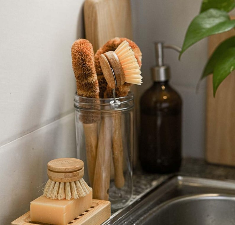 May include: A glass jar filled with four brown and beige dish brushes with wooden handles. The jar is sitting on a countertop next to a brown glass bottle with a pump dispenser.
