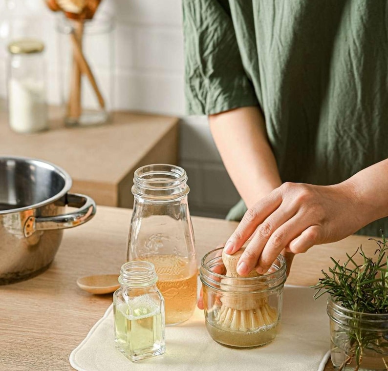 May include: A close-up of a glass jar with a wooden brush inside. The jar is sitting on a white surface with a clear glass bottle and a wooden spoon. The jar is filled with a clear liquid.